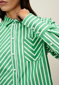 Kobieta poprawiająca kołnierzyk zielono-białej, pionowo prążkowanej koszuli z guzikami, z dopasowaną kieszenią w wzór zygzaków.