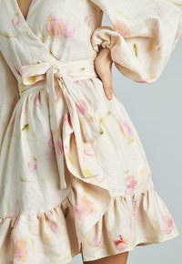 Helles beige Wickelkleid mit pastellfarbenen Blumenmustern, gerüschtem Saum und langen Puffärmeln, begleitet von einem passenden Bindegürtel. Weicher Leinenstoff.