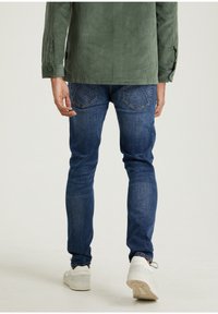 Vihreä corduroy-takki sinisten skinny-farkkujen päällä, jossa on sileä pinta ja hyvin muotoillut takataskut. Valkoiset tennarit täydentävät asua.