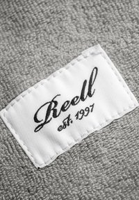 Gray textured fabric with a white label displaying the text "Reell" in black cursive and "est. 1997" in smaller font, stitched on the edge.