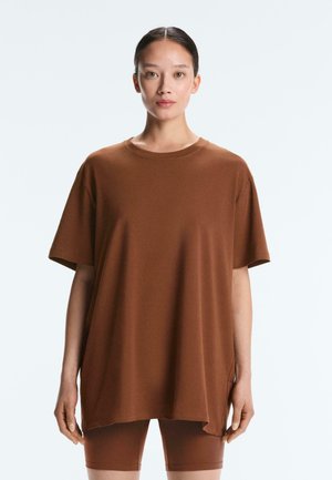 Žena s tmavými vlasmi zviazanými dozadu nosí veľké hnedé tričko a zodpovedajúce cyklistické šortky, stojaca pred jednoduchým bielym pozadím.