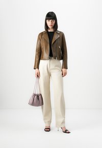 Donna che indossa una giacca di pelle marrone, una maglia nera, pantaloni beige a gamba larga, sandali neri con tacco e tiene in mano una borsa a tracolla mauve.
