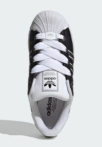 Black leather sneaker with white rubber shell toe, white laces, and three white stripes on the side. Features a textured upper and logo detail.