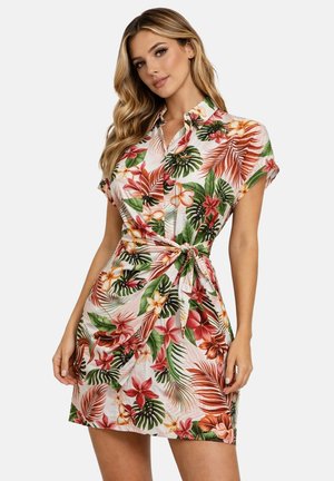 Femme portant une robe à manches courtes avec col, à motif floral tropical, nouée à la taille, posant devant un fond uni.