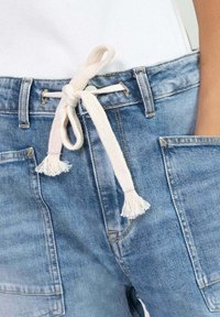 Pantalons en denim bleu avec de larges poches avant et une ceinture épaisse en tissu blanc nouée en un nœud à la taille.
