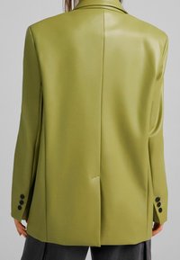Olivově zelený blejzr s hladkým, lesklým povrchem, s jedním rozventilovaným zadním dílem a čtyřmi černými knoflíky na každém rukávu. Hladká, vykrojená silueta.