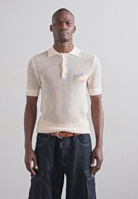 White knitted polo shirt with short sleeves, featuring a buttoned placket. Worn with dark blue cargo pants and a brown belt.