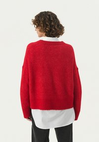 Röd oversized tröja med ribbad nederkant och mudd. Tillverkad av mjukt, strukturerat material. Lagd över en vit skjorta med krage för kontrast.