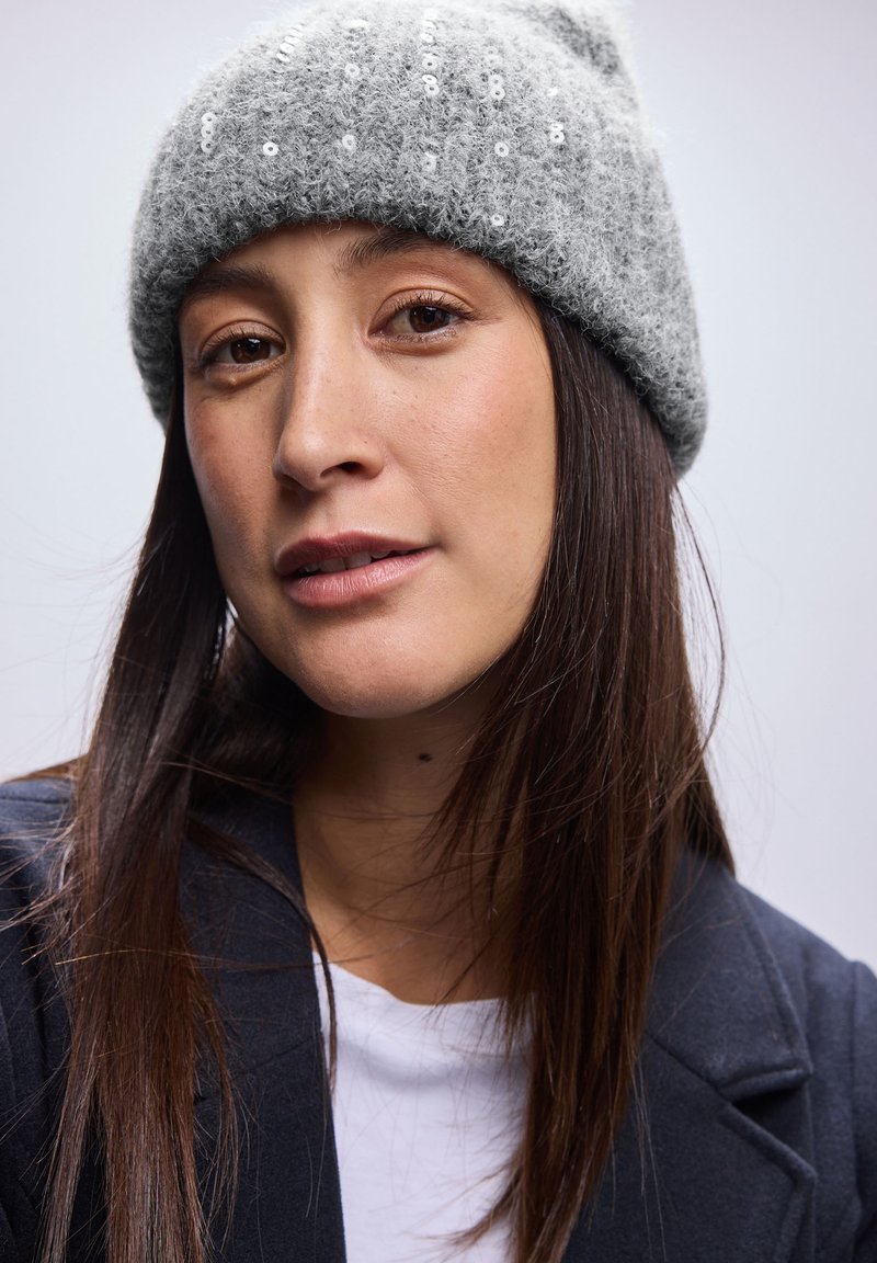 Grijze wollen muts met pailletten, lichtjes losvallend model. Model draagt een zwart colbert over een wit overhemd, gericht naar de camera.
