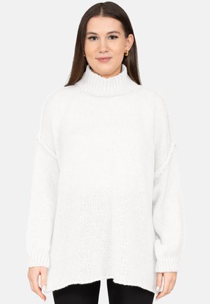 Frau trägt einen weißen Rollkragenpullover und schwarze Hosen, steht vor einem schlichten hellen Hintergrund.