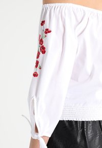 Blusa blanca sin mangas con flores rojas bordadas, cinturilla elástica y lazos en las mangas. Material de algodón ligero.