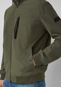 Man draagt een olijfgroene jas met ritssluiting, zij- en borstzakken, zwarte ritstrekker, geribbelde manchetten en een blauwe spijkerbroek, hand in de zak.