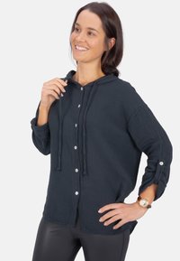 Dunkelblaue Kapuzenjacke aus strukturiertem Stoff, mit Knopfleiste, Kordeln und aufgerollten Ärmeln. Kombiniert mit glänzenden schwarzen Leggings.