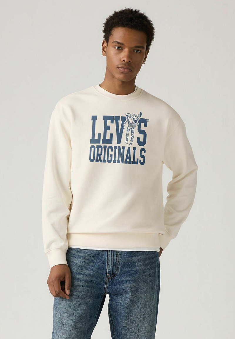 Sweat-shirt crème avec graphisme bleu "Levi's Originals". Texture douce avec poignets et ourlet côtelés, porté sur un jean. Design minimaliste.