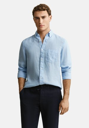 Jeune homme aux cheveux bouclés portant une chemise en lin bleu clair à manches longues retroussées et un pantalon foncé, debout avec les mains dans les poches.