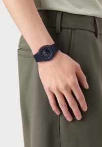 Orologio in silicone blu navy con quadrante rotondo, numeri arabi e cinturino testurizzato; indossato su una mano poggiata su un tessuto verde.