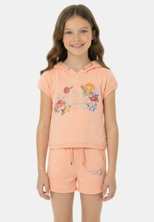 Fille souriante aux longs cheveux bruns portant un sweat à capuche à manches courtes imprimé floral pêche et un short assorti, debout devant un fond blanc.