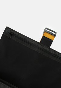 Borsa in tessuto nero con chiusura a zip. L'interno presenta una tasca a zip e una tracolla gialla con una fibbia nera. Texture liscia, forma rettangolare.