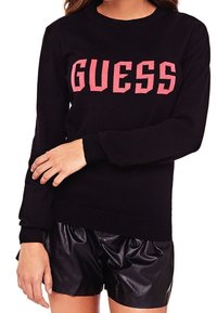 Schwarzer Pullover mit Rundhalsausschnitt und langen Ärmeln, der vorne mit einem auffälligen pinkfarbenen "GUESS"-Schriftzug versehen ist. Kombiniert mit schwarzen Shorts aus Lederimitat.