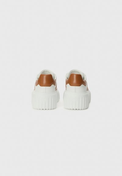 Baskets blanches avec un accent en cuir marron à l'arrière et des semelles épaisses à rainures, présentant une texture lisse et un design minimaliste.