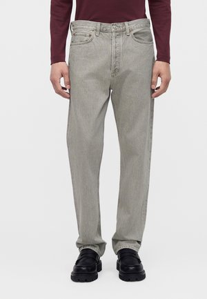 Personne portant un jean gris clair ample, une chemise bordeaux à manches longues et des chaussures noires épaisses, debout devant un fond uni.