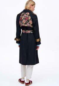 Ciemny płaszcz trencz z kwiatowym wzorem na plecach i paskiem, złote akcenty na rękawach oraz teksturowana tkanina. Luźne dopasowanie z jasnymi spodniami.