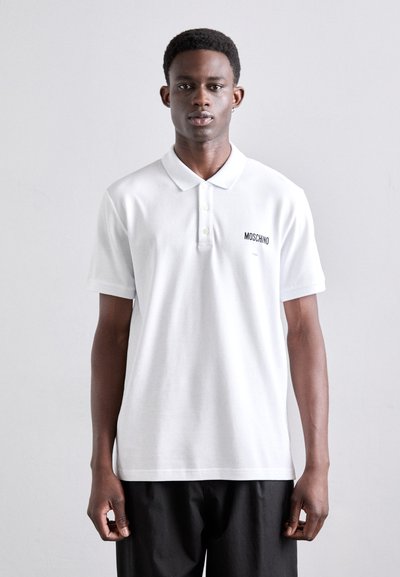 Valkoinen polo paita, joka on valmistettu sileästä puuvillasta, klassisella kauluksella ja lyhyillä hihoilla. Vasemmassa rinnassa on musta "MOSCHINO" -logo.