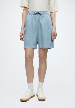 Persoon draagt lichtblauwe knielange broek met trekkoord, witte sokken, witte sneakers en een beige top met korte mouwen.