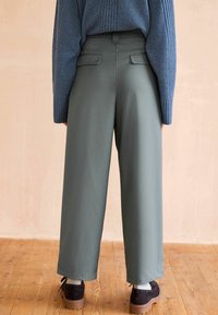Pantaloni a gamba larga in tessuto verde tenue, con tasche laterali e design sartoriale, abbinati a stivali neri alla caviglia su un pavimento di legno.