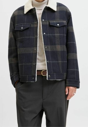 Man draagt een donker geruit jack met witte fleece kraag over een crèmekleurige coltrui, donkere broek en een bruine riem, handen deels in de zakken.