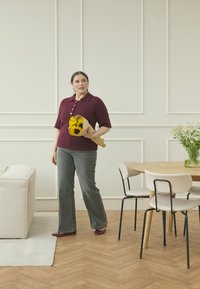 Bordeaux gebreide polo sweater, grijze uitlopende broek, met een boeket zonnebloemen, in een lichte kamer met een ronde houten tafel en moderne stoelen.