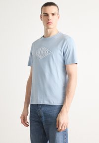Lyseblå bomull t-skjorte med korte ermer, med en hevet diamantlogo i lys grått i sentrum. Bært med blå jeans.