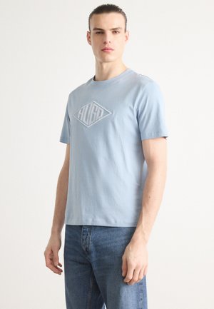 Light blue cotton t-shirt with short sleeves, featuring a raised diamond logo in light gray at the center. Worn with blue jeans.