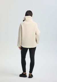 Cremefarbene Fleecejacke mit hohem Kragen, lockerer Passform und strukturierter Oberfläche. Kombiniert mit schwarzen Leggings und Schuhwerk.