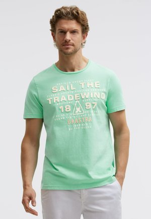 Mann mit kurzen lockigen Haaren, der ein mintgrünes T-Shirt mit beigem nautischem Schriftzug "Sail the Tradewind 1897 Gaastra" und weiße Hosen trägt.