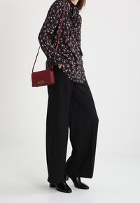 Blusa nera floreale con cravattino, abbinata a pantaloni larghi neri e una borsa a tracolla bordeaux. Stivali neri con punta a punta completano il look.