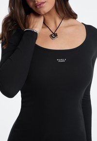 Schwarzes, geripptes Longsleeve-Top mit Rundhalsausschnitt, das das Logo "Guess Jeans" zeigt. Accessoires sind ein silbernes Armband und eine herzförmige Anhängerkette.