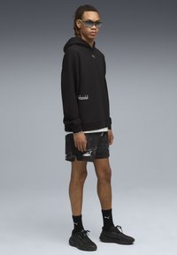 Ung mand iført sort Puma hættetrøje, mønstrede sorte shorts, sorte Puma strømper, sorte sneakers og futuristiske solbriller, stående mod en grå baggrund.