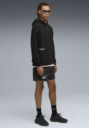 Ung mand iført sort Puma hættetrøje, mønstrede sorte shorts, sorte Puma strømper, sorte sneakers og futuristiske solbriller, stående mod en grå baggrund.