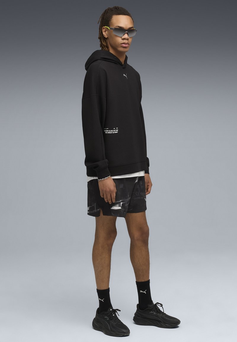 Ung mand iført sort Puma hættetrøje, mønstrede sorte shorts, sorte Puma strømper, sorte sneakers og futuristiske solbriller, stående mod en grå baggrund.