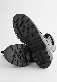 Semelles de bottes noires présentant un motif de semelle robuste, en matériau en caoutchouc texturé, avec la marque 'NEXT' embossée sur le côté.