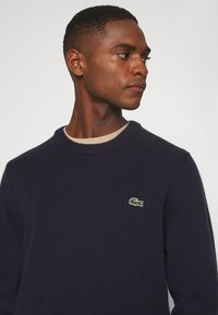 Jersey de punto azul marino con cuello redondo y un pequeño logo del cocodrilo Lacoste en el pecho, fabricado con material suave y texturizado.