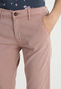 Světle růžové bavlněné kalhoty s přední kapsou, zapínáním na knoflík a texturovaným povrchem. Detailní pohled na oblast pasu a nohou.