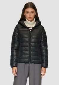 Doudoune noire matelassée avec une fermeture éclair à l'avant, une capuche et une texture matelassée. Dotée de poches latérales et d'une silhouette ajustée, portée par-dessus un col roulé crème.