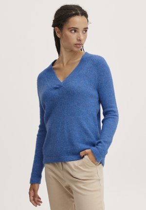 Pull bleu à col en V en matériau doux, avec des manches longues et une coupe décontractée. Assorti avec un pantalon beige.