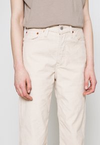 Pantalon en velours côtelé beige avec une coupe droite, doté d'une fermeture par bouton et fermeture éclair à l'avant, de deux poches avant, et d'une texture côtelée subtile.
