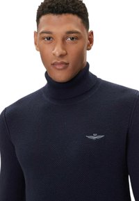 Maglione a collo alto lavorato a maglia color blu navy con motivo testurizzato e logo argentato sul lato sinistro del petto. Tessuto liscio ed elastico, design aderente.