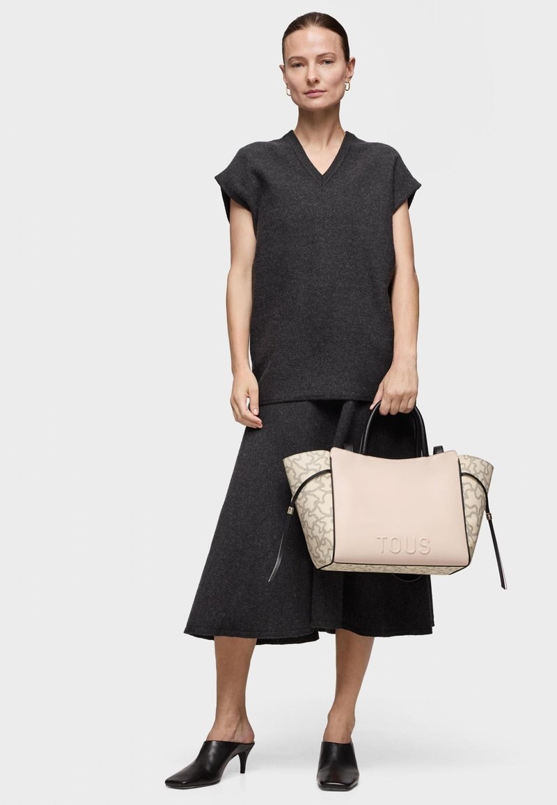 Top de punto gris con cuello en V combinado con una falda gris, sosteniendo un bolso tote beige y estampado con "TOUS" grabado en el costado. Lleva usados unos zuecos negros.