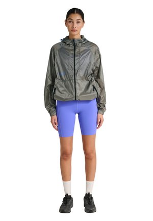 Vrouw die een grijze windjack met capuchon draagt, felpaarse fietsshorts, witte sokken en zwarte sneakers, staand met het gezicht naar voren gericht.