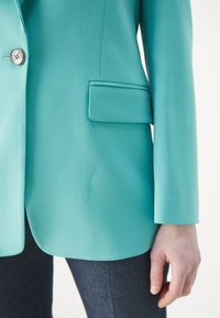 Tyrkysový blejzr s hladkou texturou, jedním stříbrným knoflíkem, přiléhavým střihem a boční kapsou, spárovaný s tmavými džíny.